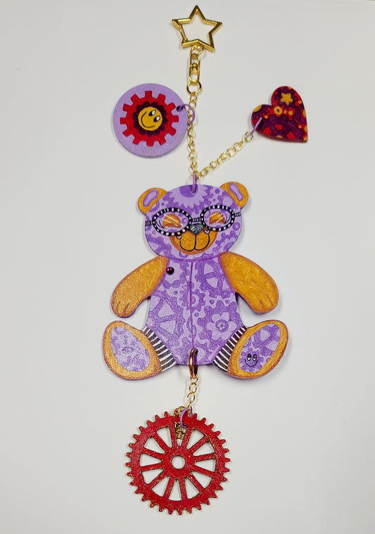 GEAR BEAR – Bibli

Meet Bibli, the regal GEAR BEAR of wisdom and wonder. Painted in rich purples, deep reds, and glowing golds, Bibli evokes ancient libraries, hidden stories, and magical realms waiting to be discovered. Her swirling gears represent knowledge in motion, while her glittered back gleams like a gilded book cover. Crystal accents add a touch of opulence fit for a queen of curiosity.

Each GEAR BEAR comes with three accessory charms—including a&nbsp;signed, hand-painted Geartopia gear tag by art